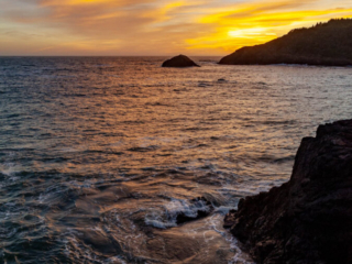 The ocean roils around some rocks below a cloudy sunset