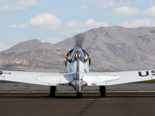An airplane sits on the tarmac idling. A person sits inside the cockpit and holds their hands outside of the window.