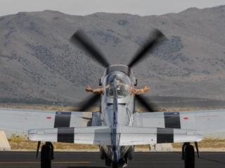 An airplane sits on the tarmac idling. A person sits inside the cockpit and holds their hands outside of the window.