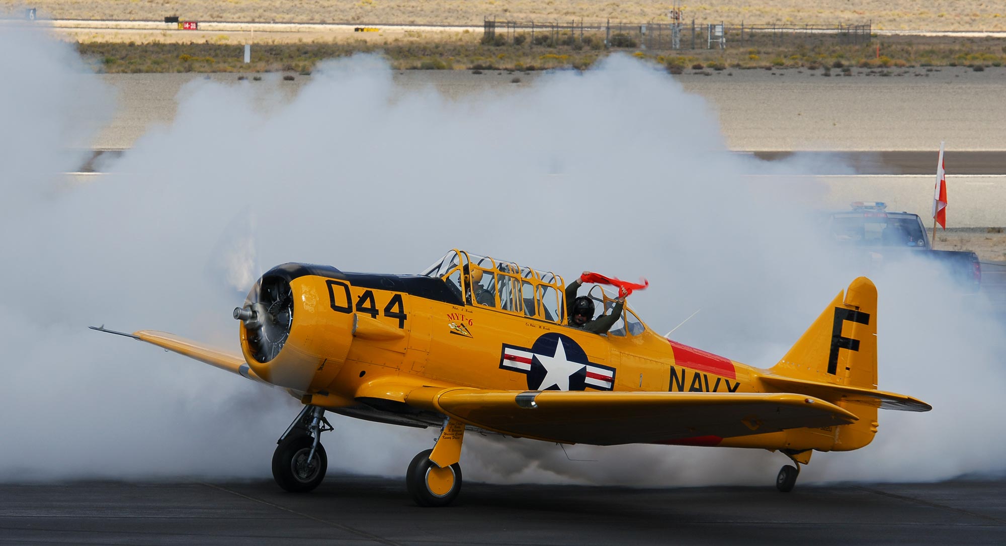 An airplane taxies across the tarmac and blows smoke in celebration. A person leans out of the back window and waves an orange flag.