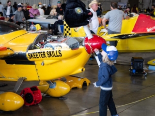 An airplane parked in a pit area has a mask on the propeller. A child looks up at the mask.