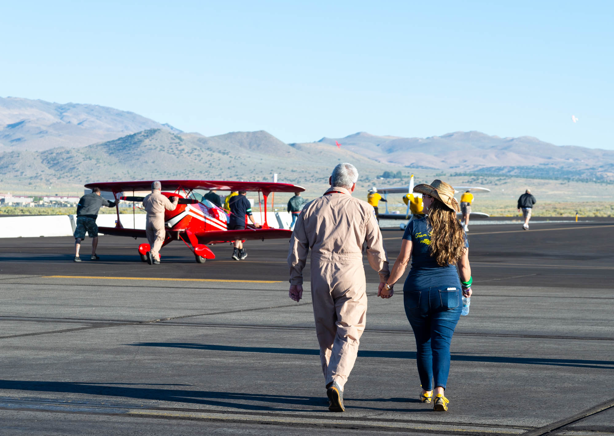 A man in a flight suit holds hands with a woman while they walk towards two biplanes.