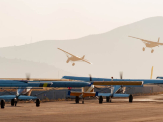 A number of STOL planes sit idling on the ramp at golden hour. Two STOL airplanes race against each other in the background, seen over the top of the idling planes.