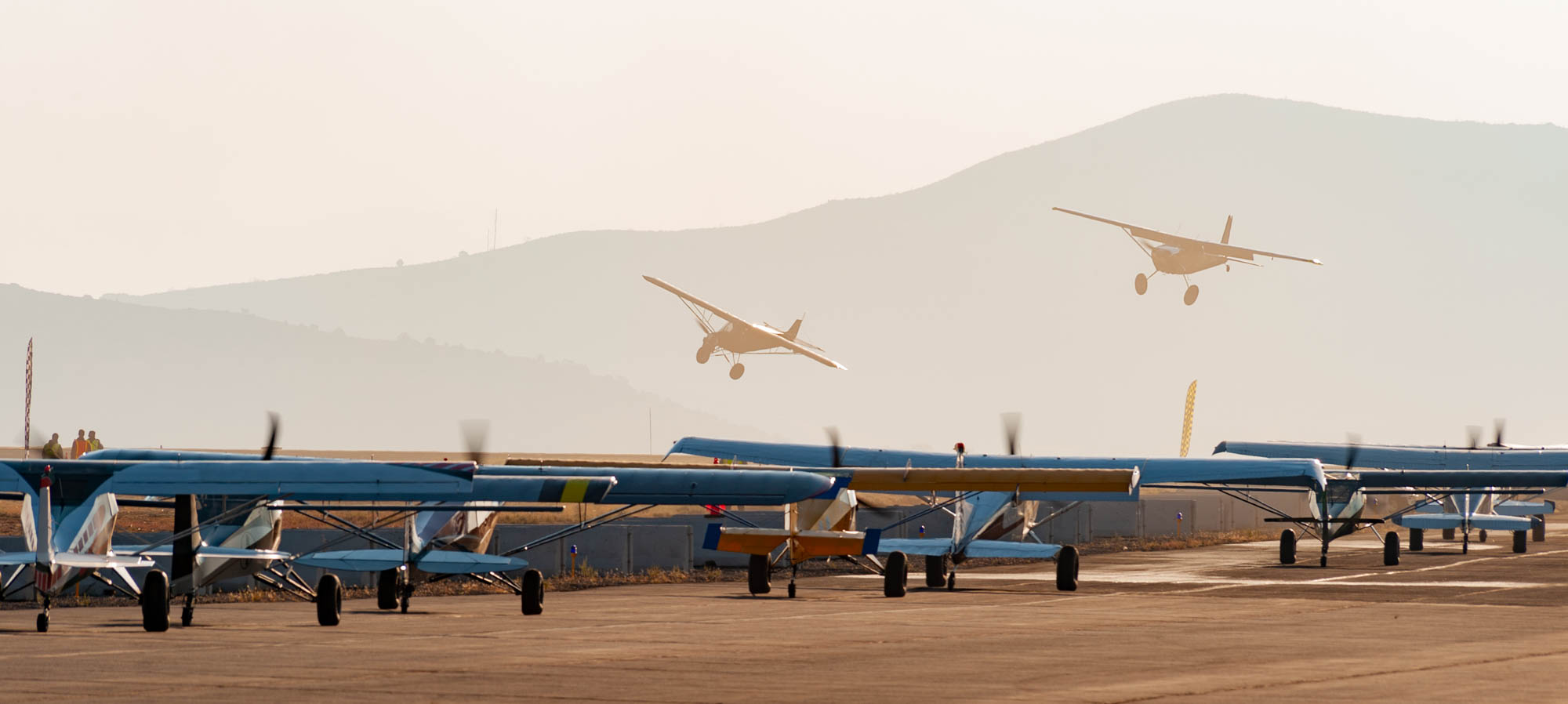 A number of STOL planes sit idling on the ramp at golden hour. Two STOL airplanes race against each other in the background, seen over the top of the idling planes.