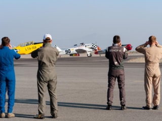 Four men in flight suits stand facing away from the camera covering their ears while airplanes idle nearby.