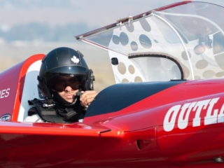 A man sits in flight gear in the cockpit of an airplane and looks intently at the camera