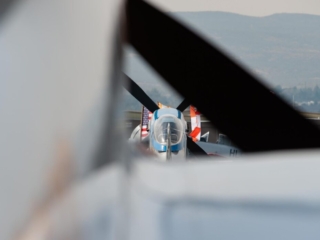 A photo of an airplane and some flags, as seen through the wing and propeller of another airplane.