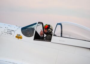 A man sits in the cockpit of an airplane and gives a hand signal to someone out of frame.