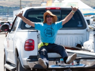 A man in a wide hat sits in the back of a pickup, giving two shaka signs