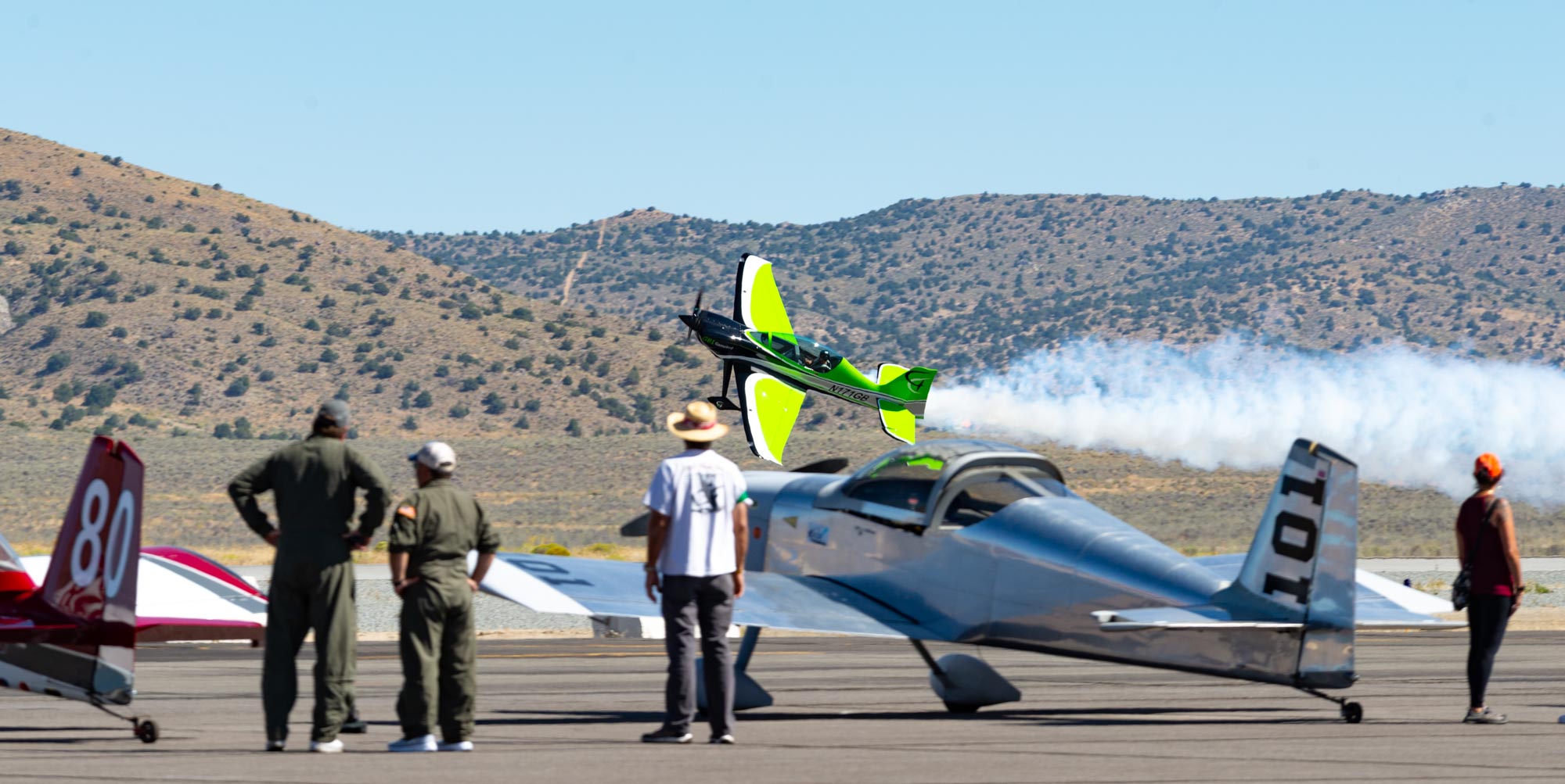 A bright green airplane flies dramatically past a group of parked airplanes. The airplane is flying very low and at a sharp angle, and has show smoke coming out of it.