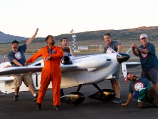 A group of people stand around their airplane, making goofy poses. The Reno Air Races home pylon is in the background