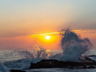 Some waves explode off of some rocks in front of a sunset.