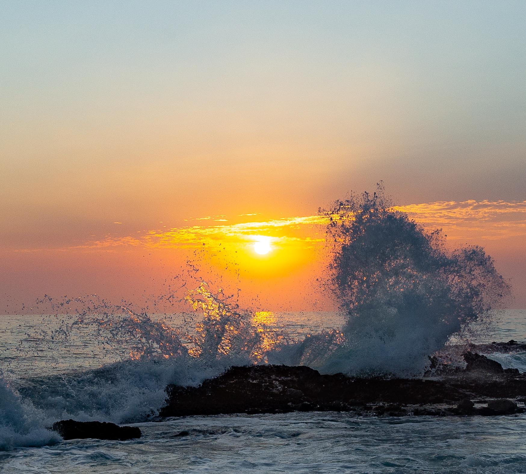 Some waves explode off of some rocks in front of a sunset.