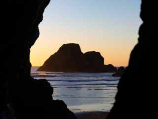 A rocky island in the ocean is framed by two large rocks. The sky is clear and the sun is setting.