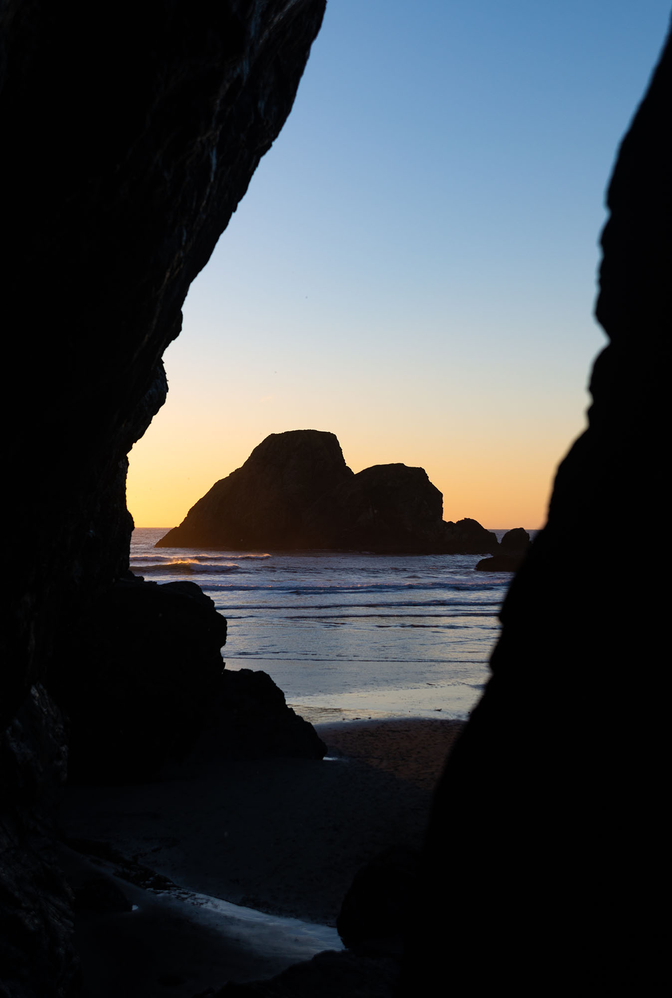 A rocky island in the ocean is framed by two large rocks. The sky is clear and the sun is setting.