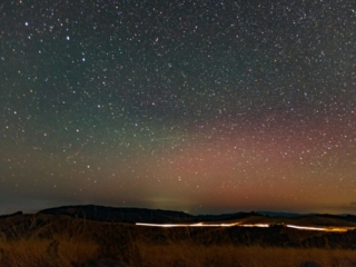 stars, shooting stars, and the northern lights sit over some grass and a streak of car's headlights
