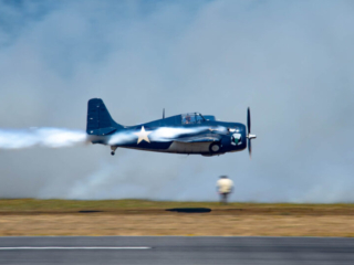 An airplane flies low over the runway, with performer smoke coming from the engine