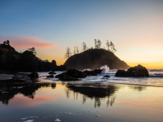 Ocean waves crash on some rocks. The sand in the foreground is well-wetted, reflecting the rocks, trees, and bright sunset in the background